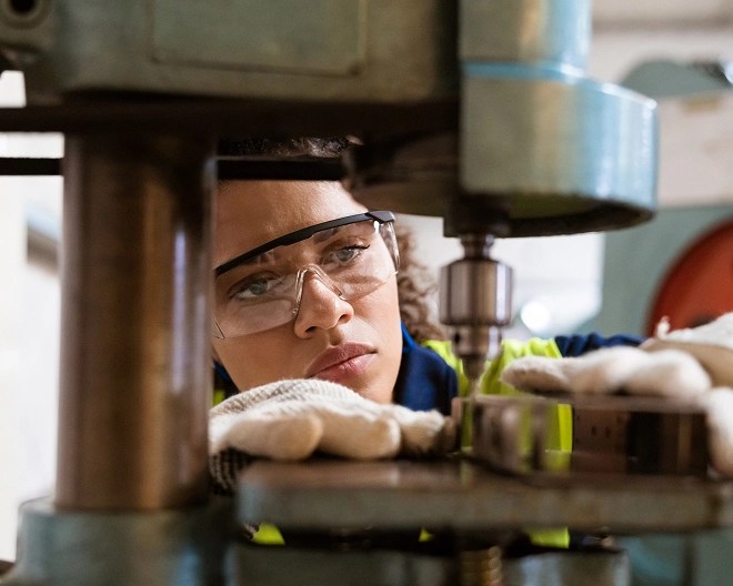 Worker in high-visibility gear and gloves operates a drill press in an industrial setting, emphasizing safety and precision in machinery use.