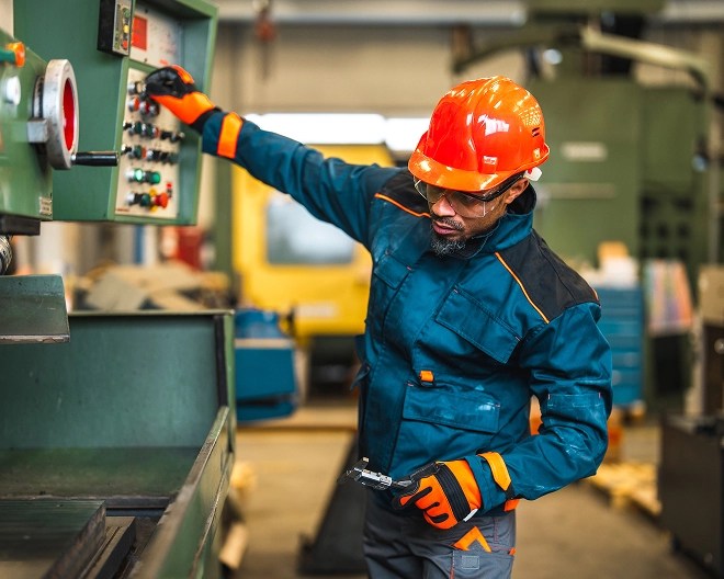 Industrial worker in safety gear operates a control panel on heavy machinery in a factory setting.