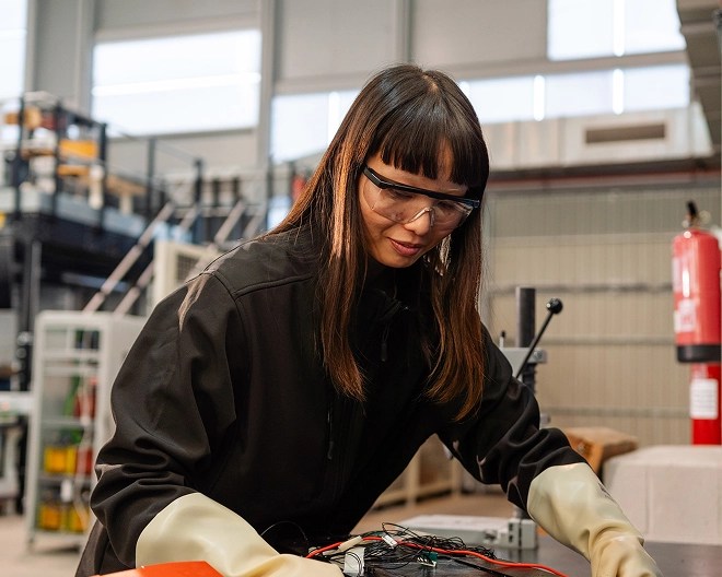 Technician in protective gloves and dark jacket works on a wired electronic device at a table in a workshop, surrounded by industrial equipment and shelves.