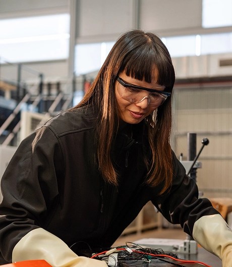 Technician in protective gloves and dark jacket works on a wired electronic device at a table in a workshop, surrounded by industrial equipment and shelves.