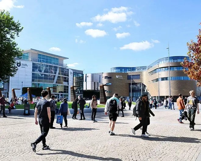 Students walk and gather in a sunny courtyard on Teesside University’s campus. Modern buildings labeled “University Library” and “University Student Services” are in the background.