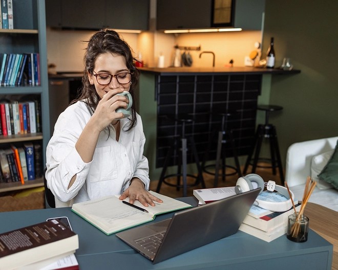 Person sitting at a desk in a cozy home workspace with a laptop, books, notebook, and headphones; a bookshelf and kitchen are visible in the background.