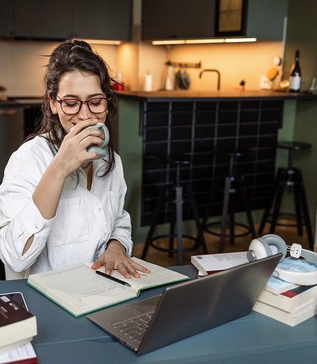 Person sitting at a desk in a cozy home workspace with a laptop, books, notebook, and headphones; a bookshelf and kitchen are visible in the background.