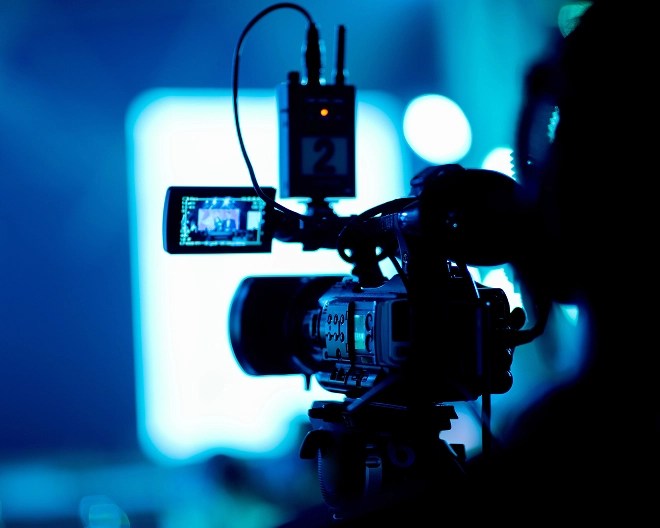 Professional video camera set up with external monitor and microphone, filming in a studio with a blue-lit background.