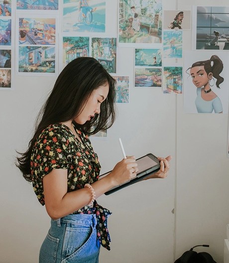 Person using a tablet and stylus in a creative workspace decorated with vibrant illustrations, plants, and digital screens.