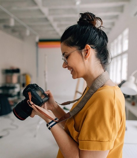 Person holding a DSLR camera in a bright studio space with desks, chairs, and large windows.
