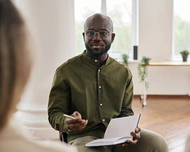 Male therapist in a dark green button-up and glasses addresses a client as he takes notes.
