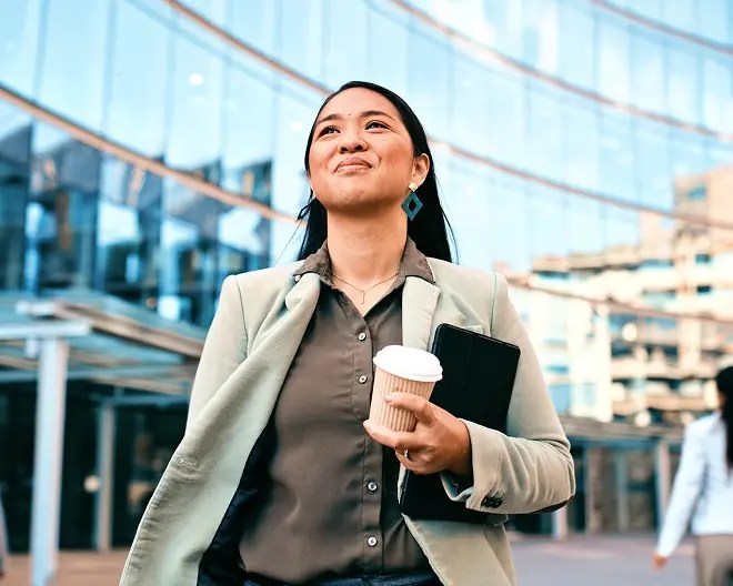 A hopeful businesswoman walks to work, holding a cup of coffee and a tablet, gazing upward with modern office buildings towering in the background.