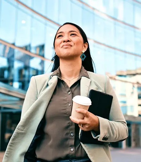 A hopeful businesswoman walks to work, holding a cup of coffee and a tablet, gazing upward with modern office buildings towering in the background.