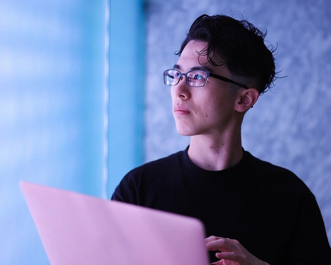 Person in a black shirt and glasses using a pink laptop while gazing out a window.