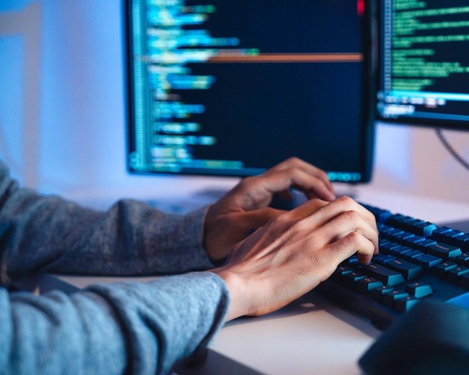 Person typing on a mechanical keyboard in a dimly lit room with blue lighting, surrounded by dual monitors displaying code.