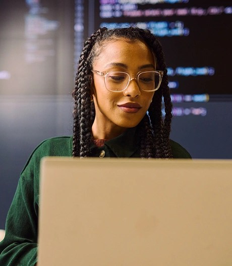 Person with braided hair and glasses working on a laptop in front of large screens filled with code.