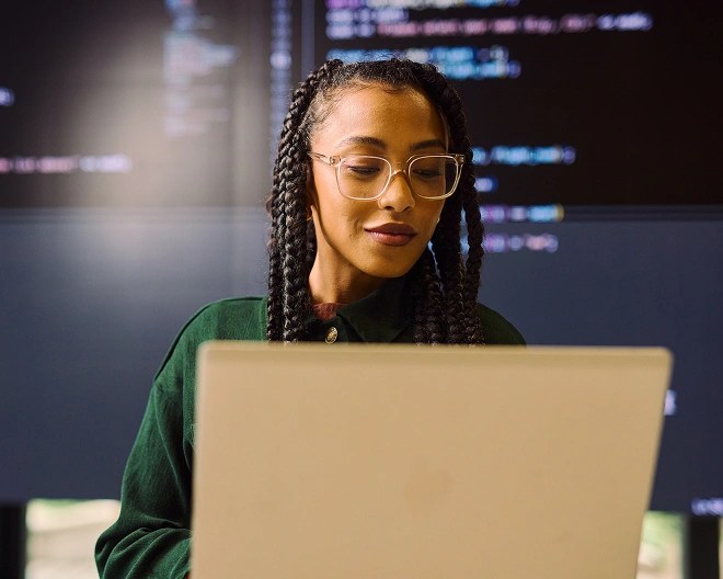 Person with braided hair and glasses working on a laptop in front of large screens filled with code.