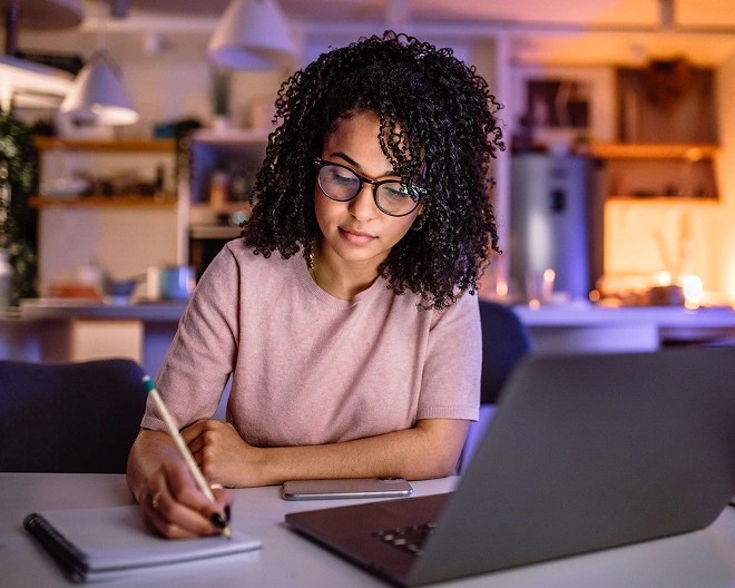 Person with curly hair and glasses working on a laptop and writing in a notebook at a kitchen table