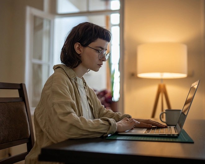 Person in a light shirt and glasses working on a laptop at a dark table, with a cup nearby.