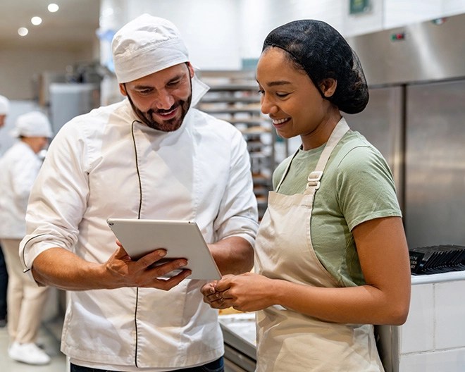 Chef in white uniform showing something on a tablet to a smiling colleague in a beige apron.