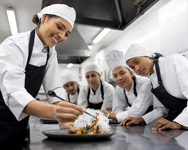 Chef carefully plating a gourmet dish while four others observe in a commercial kitchen.