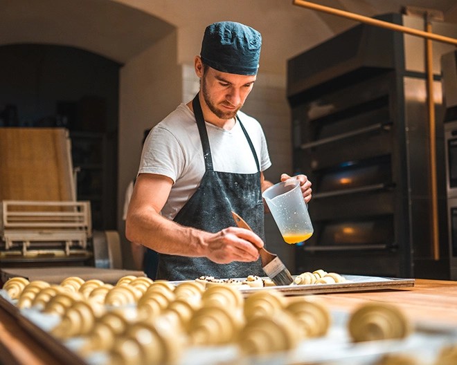 Person in a black apron brushing egg wash onto pastries in a commercial bakery.
