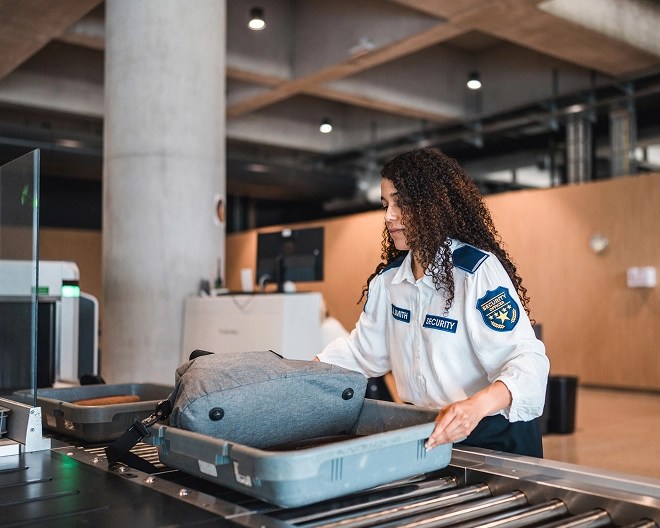 Airport security officer inspecting a passenger’s bag at an X-ray scanner checkpoint.