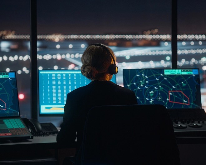 Air traffic controller wearing a headset monitors multiple screens displaying flight paths and radar data in a control tower overlooking a lit airport runway at night.