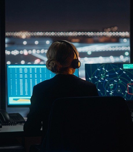 Air traffic controller wearing a headset monitors multiple screens displaying flight paths and radar data in a control tower overlooking a lit airport runway at night.