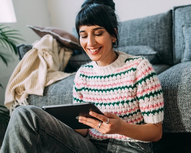 Person in a striped sweater and gray jeans sits on the floor in front of a couch, smiling while using a tablet in a naturally lit living room with plants and soft furnishings.
