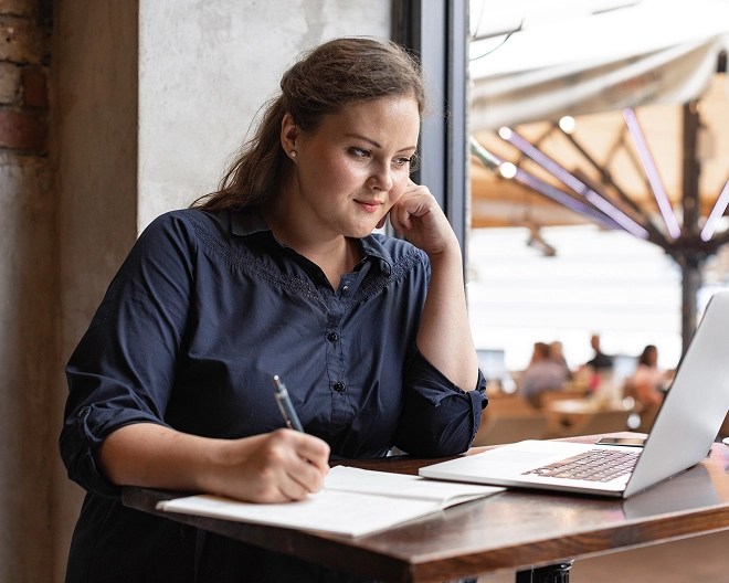 Individual in a dark blue shirt writes in a notebook while referencing a laptop at a café table near a window, with other patrons visible in the softly lit background.