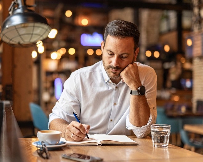 Person in a white shirt writes in a notebook at a warmly lit café table with coffee, water, and a smartphone, resting their head on one hand in a thoughtful pose.