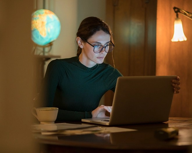 Person in glasses and a dark green top works on a laptop at a desk with a lamp, papers, and a globe, suggesting a studious or remote work environment.