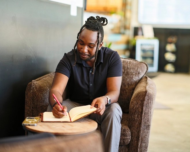 Person writing in a notebook at a small round table in a cozy indoor setting, with a drink nearby.