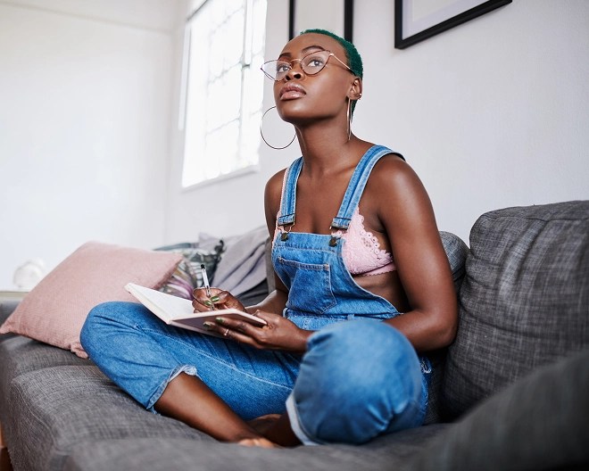 Person wearing overalls and glasses, lost in thought as she writes in a notebook on a grey couch in a sunlit room.