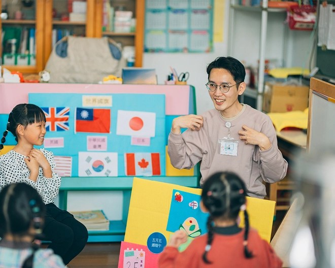 Teacher engaging with young students in a classroom decorated with international flags and colourful crafts.