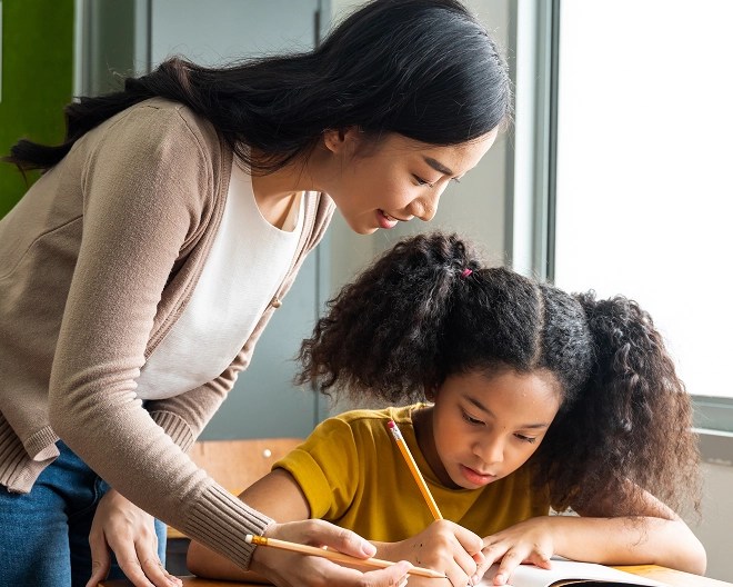 Adult assisting a young student with writing in a sunlit classroom setting.