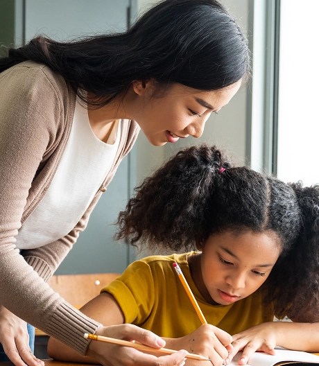 Adult assisting a young student with writing in a sunlit classroom setting.
