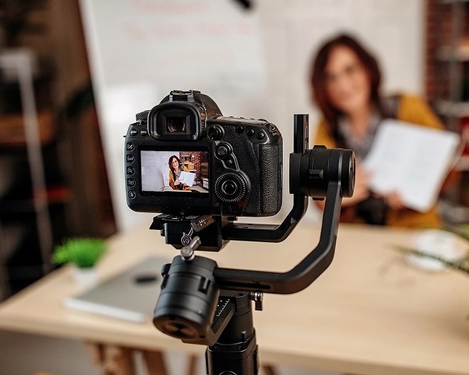 Professional camera on a gimbal tripod records a person holding up a paper at a desk, with the camera screen in sharp focus and a whiteboard in the background.
