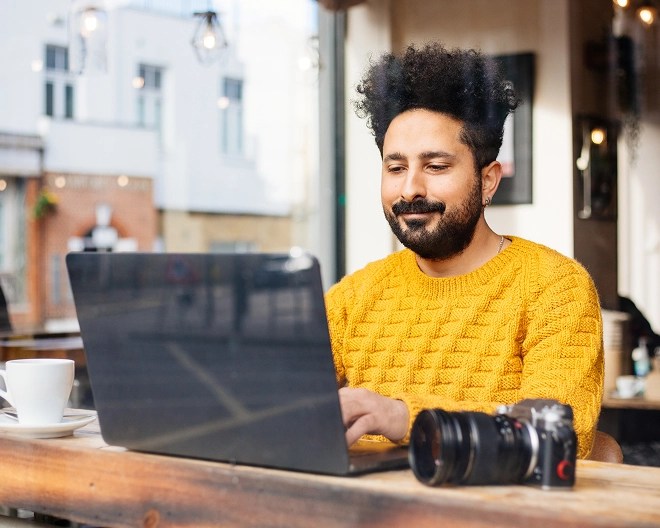 Person in a bright yellow sweater works on a laptop at a café table with a coffee cup and camera, surrounded by natural light and large windows.