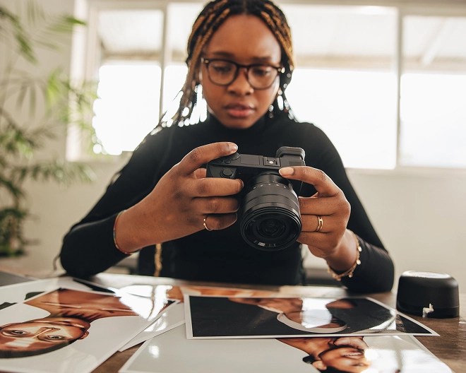Photographer in glasses examines a digital camera at a table covered with printed portrait photos, in a well-lit indoor space with plants and large windows.