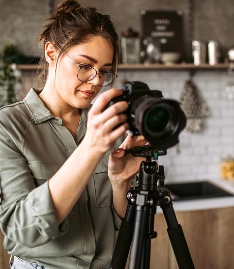 Person in a green shirt adjusts a camera on a tripod in a modern kitchen with white tiles, wooden cabinets, and cooking tools, preparing for a shoot.