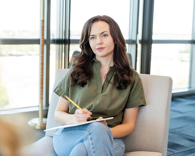 Individual with long brown hair writes in a notepad with a yellow pencil while seated in a bright room with large windows and a floor lamp.