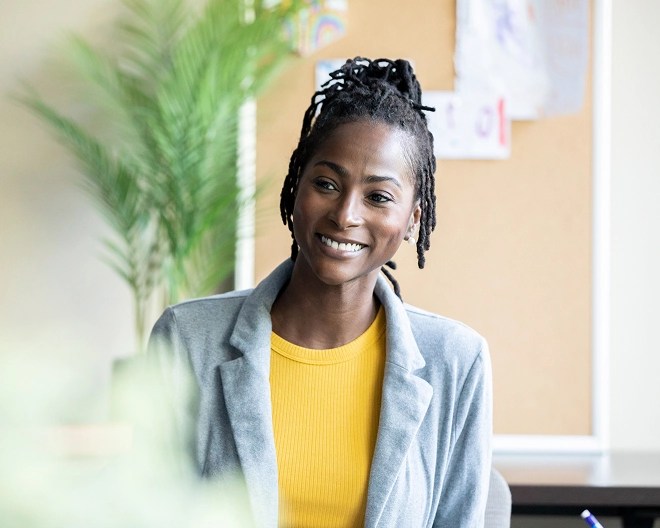 Person in a light gray blazer and mustard yellow top smiles in a well-lit office with a corkboard and green plant in the background.