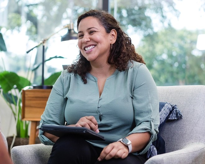 Person in a light green blouse sits on a couch holding a tablet, smiling in a bright room with large windows, plants, and a side table, suggesting a relaxed professional conversation.