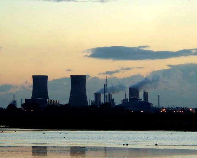 Industrial facility with cooling towers and chimneys emitting smoke, located near a body of water under a partly cloudy sky at sunrise or sunset.