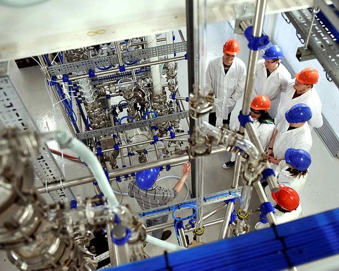 Group of people in lab coats and hard hats gathered around a network of metal pipes, valves, and gauges in an industrial training facility.