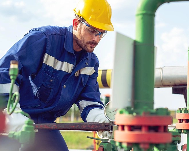 Person in a yellow hard hat and blue uniform adjusting a valve on outdoor industrial piping with additional equipment in the background.