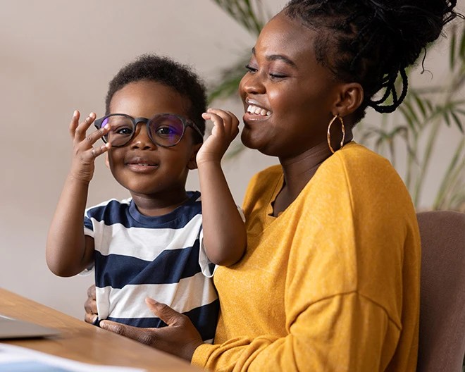 Adult in a mustard yellow top holding a child in a striped shirt who is playfully wearing oversized eyeglasses, indoors with a plant and neutral wall in the background.
