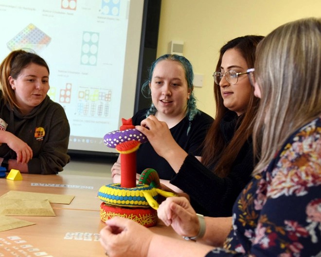 Four people sitting around a table interacting with a colourful tactile object, with a projection screen in the background displaying diagrams and numbers in a classroom setting.