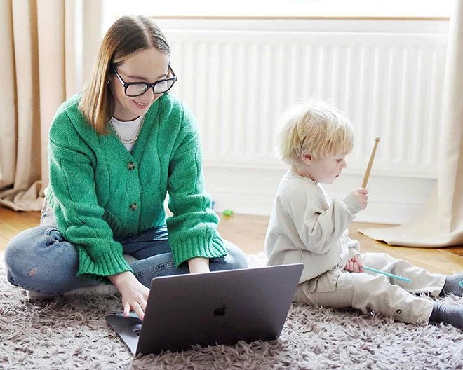 Adult in a green sweater using a laptop while sitting on the floor next to a child holding a wooden stick, in a cozy room with a radiator and beige curtains.
