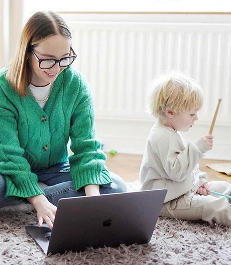 Adult in a green sweater using a laptop while sitting on the floor next to a child holding a wooden stick, in a cozy room with a radiator and beige curtains.