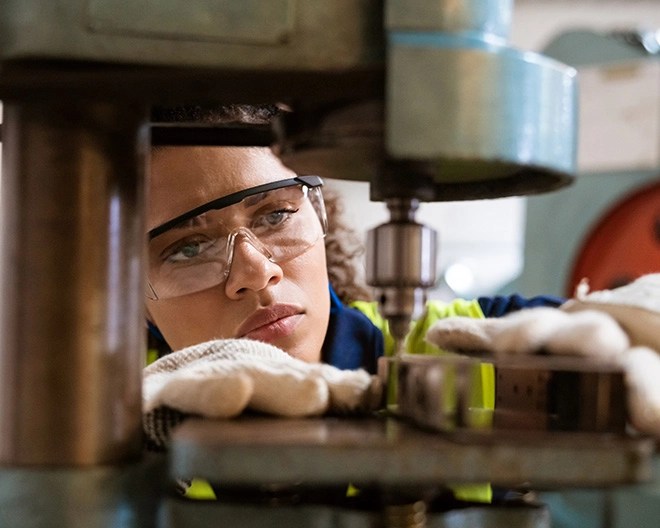 Individual wearing safety glasses and gloves operating a drill press in a workshop, positioning a metal workpiece under the tool head.
