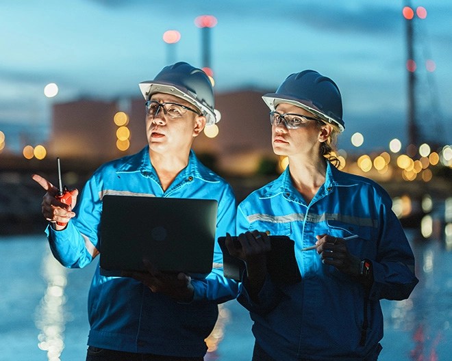 Two people in blue protective uniforms and safety helmets standing near a body of water at night, using a laptop and tablet with an industrial facility in the background.